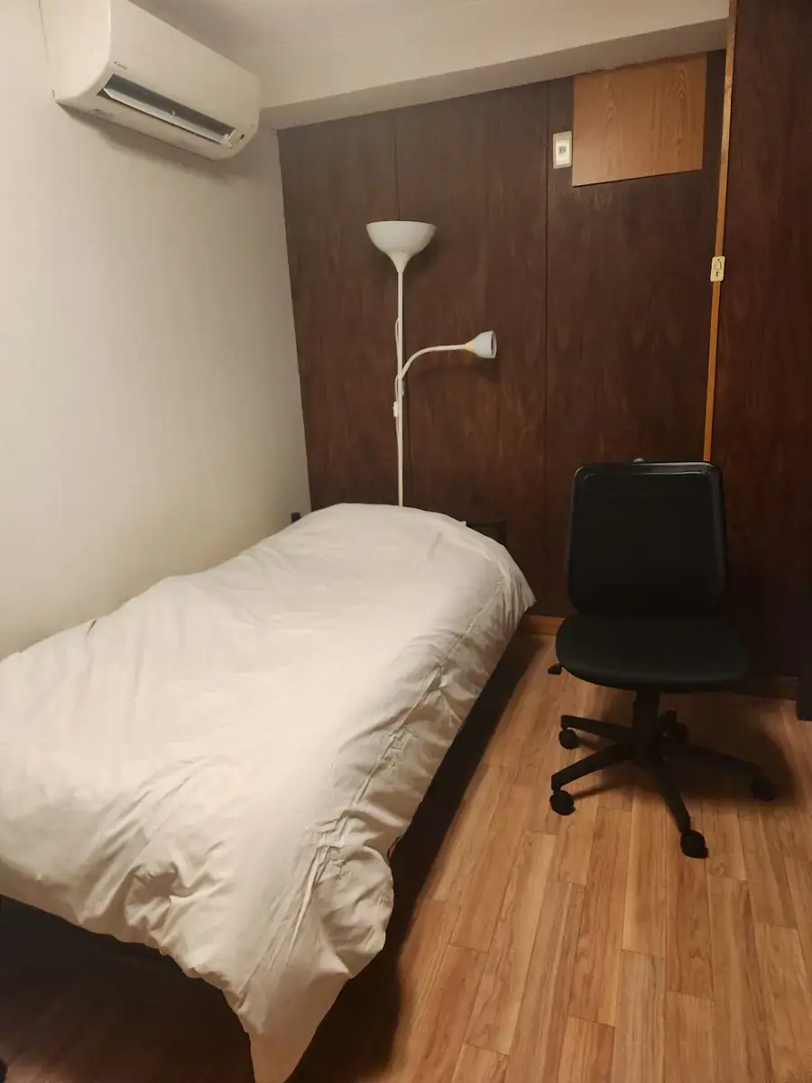 셰어하우스 방. 침대와 사무용 의자, 스탠드 조명이 놓여 있다.
